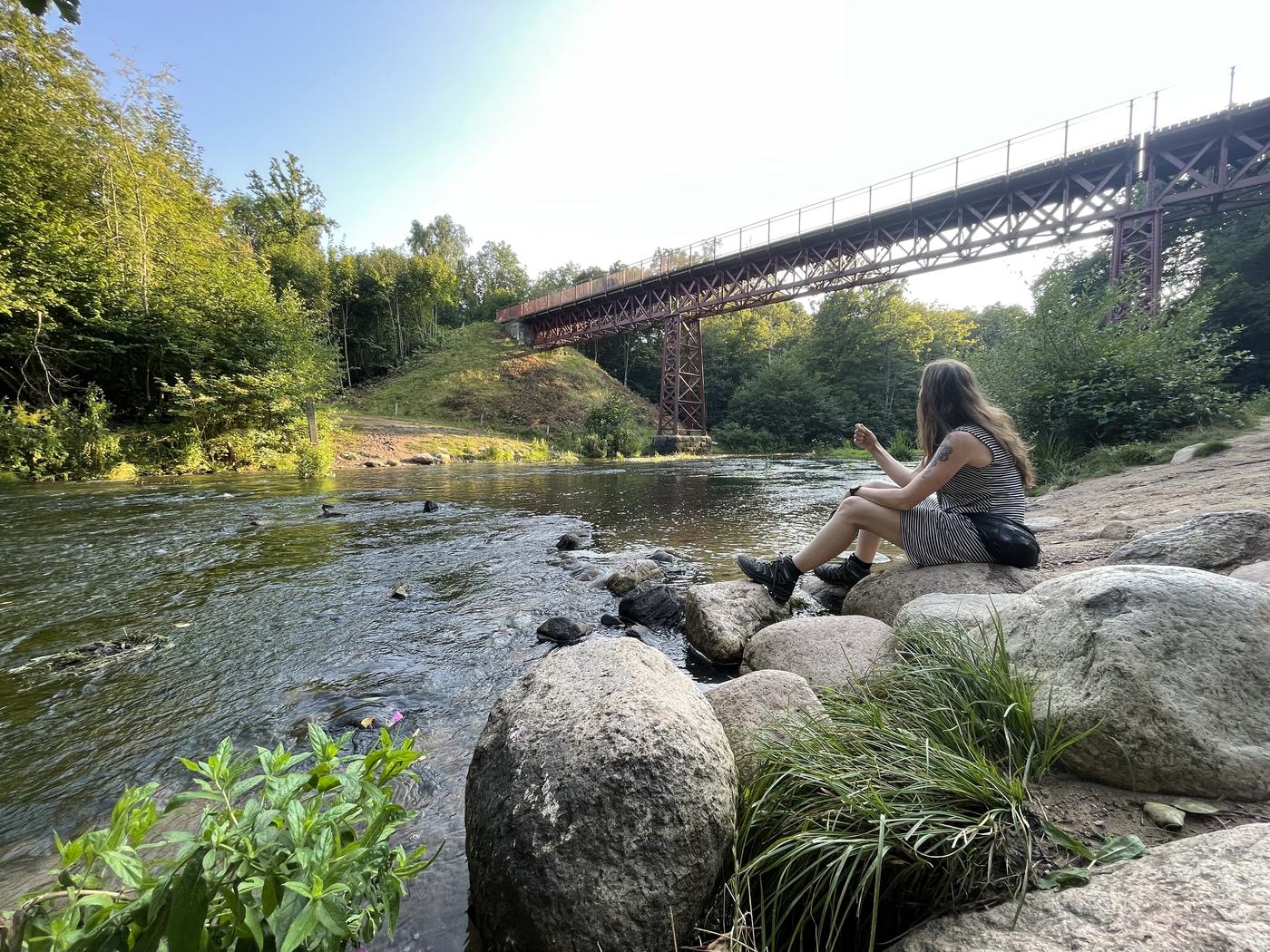 Maja sidder på store sten ved en å i aftensol med en gammel jernbanebro i baggrunden